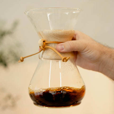 Hand holding a glass coffee maker with a paper filter and coffee inside, blurred background