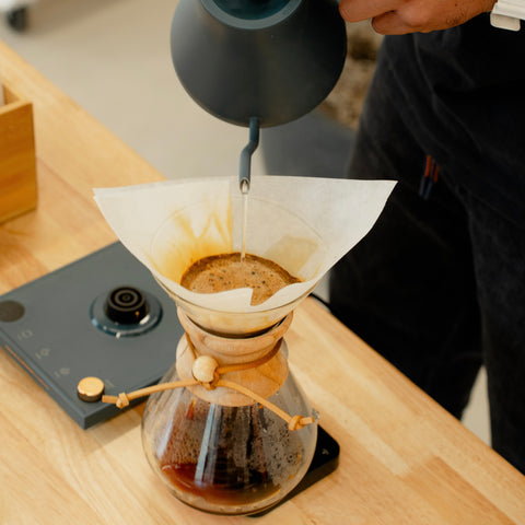 Person pouring water into a Chemex filter on a wooden counter.